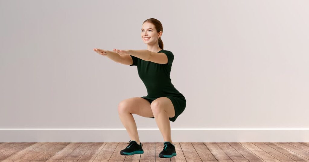A young fit woman performing a bodyweight squat indoors, demonstrating proper leg workout form without equipment