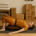 Woman in Iyengar Yoga studio performing Setu Bandha Sarvangasana (Bridge Pose) on a bolster with belt support, surrounded by props.